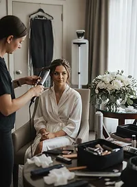 A stylist curling a bride's hair while she sits in a robe in a hotel suite, a white rose bouquet on the table beside her