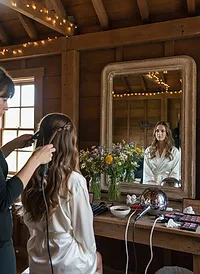 A stylist finishing a bride's half-up braid and waves in a rustic barn, with wildflowers and string lights around a wooden mirror