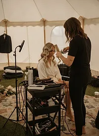 A stylist shaping soft waves in a bride's blonde hair at a mobile station inside a white tent