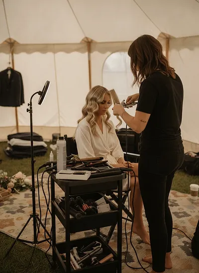 A stylist shaping soft waves in a bride's blonde hair at a mobile station inside a white tent
