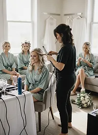 A stylist curling a bride's hair as bridesmaids in sage robes wait on the couch in a bright getting-ready room