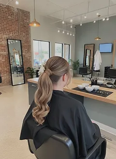 A woman at a styling station seen from behind, her hair in a braided crown updo with soft loose pieces