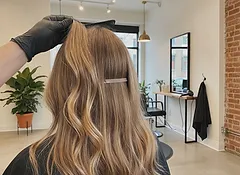 A client in a black cape from behind with long dark-blonde waves, a stylist's hand holding the ends to show the added length