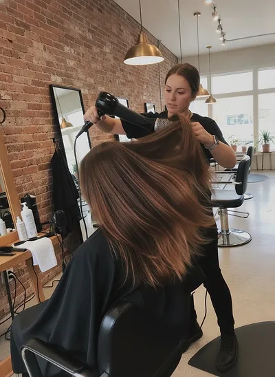 A stylist blow-drying a client's long brown hair with a round brush